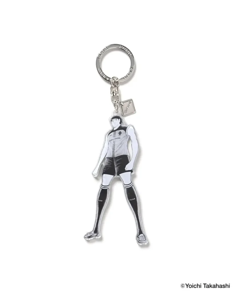 Minimal acrylic-style keychain accessory featuring a manga-inspired football player illustration by Yoichi Takahashi for F.C. Real Bristol. The monochrome character is depicted in a sleeveless football kit, knee-high socks, and cleats with subtle Bristol branding, attached to a polished silver key ring and metal tag. The collectible design merges Japanese manga aesthetics with football culture and contemporary streetwear merchandising