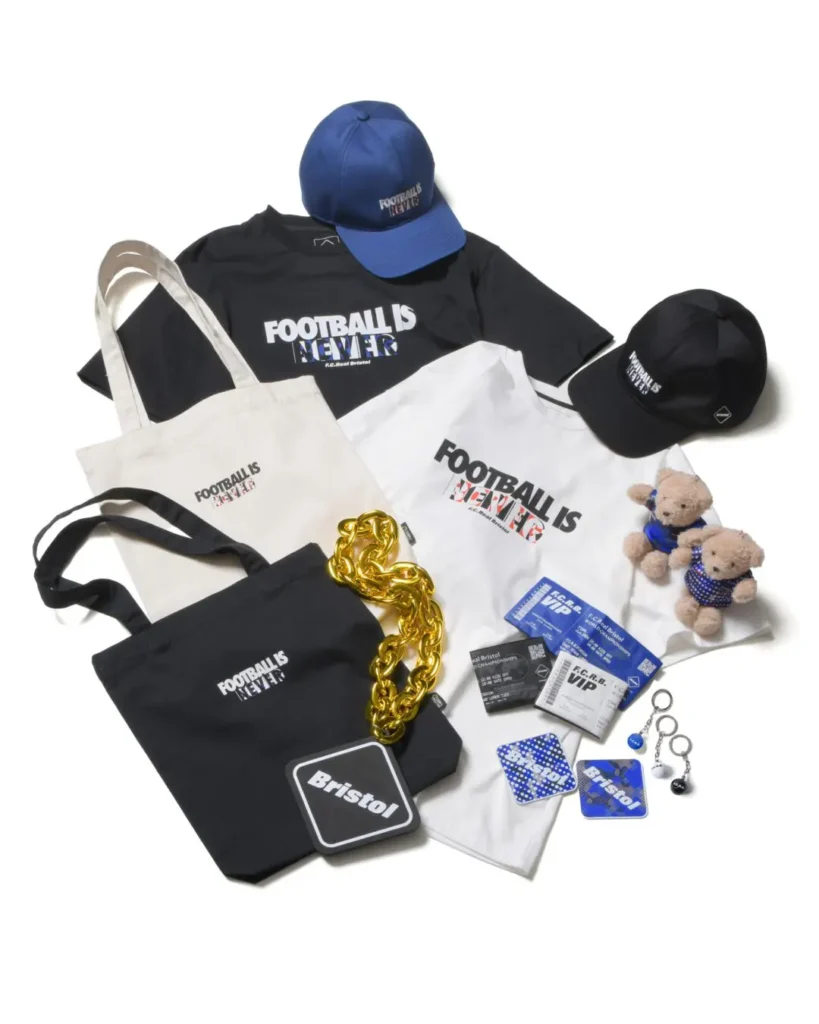 Flat-lay presentation of SOPH. Co., Ltd.’s “FOOTBALL IS NEVER” merchandise capsule arranged on a clean white background. The collection includes black and white graphic T-shirts, embroidered blue and black caps, canvas tote bags, teddy bear accessories dressed in blue football-inspired outfits, VIP-style passes, keychains, and branded lifestyle accessories. Bold monochrome branding is contrasted by electric blue accents and metallic gold chain detailing, reinforcing the campaign’s fusion of football culture and contemporary streetwear design