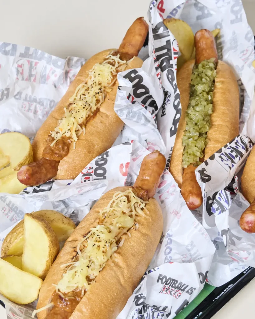 Assorted gourmet hot dogs served in toasted buns atop custom “FOOTBALL IS NEVER” branded paper liners. The spread includes sausages topped with melted shredded cheese, rich chili-style sauce, and finely chopped relish, accompanied by thick-cut potato wedges. The casual presentation blends stadium snack culture with elevated café styling, reinforcing the football-inspired lifestyle aesthetic associated with SOPH. and F.C. Real Bristol branding