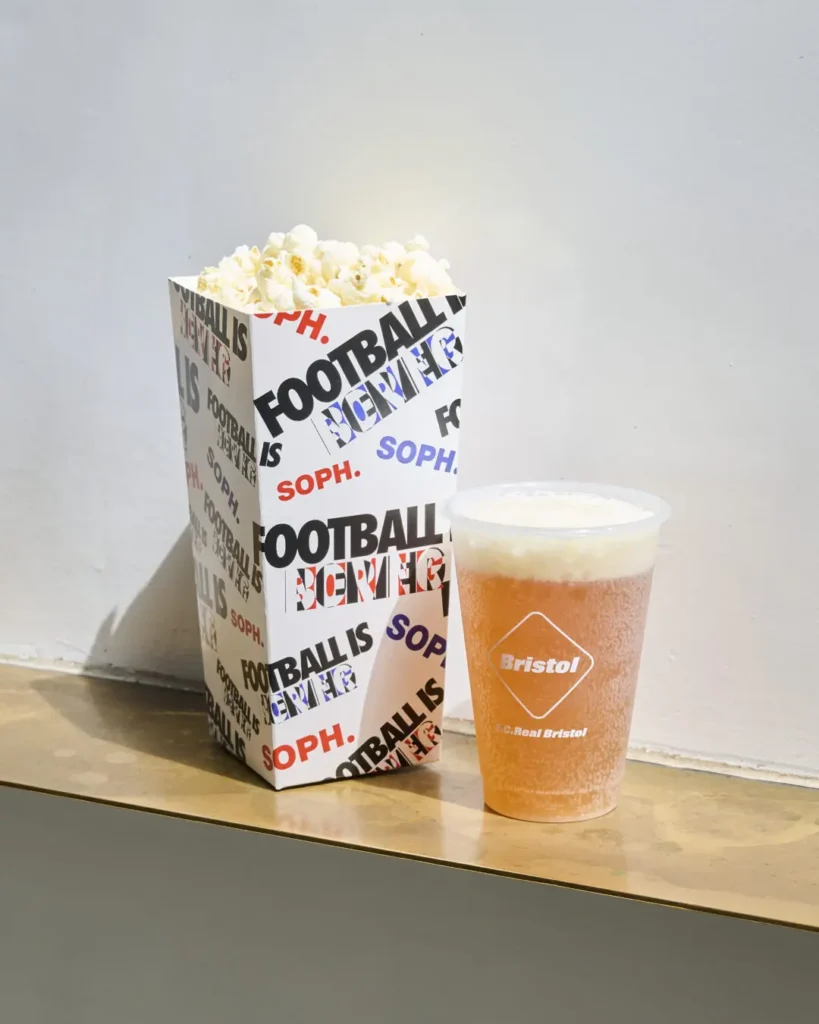 Minimal football-inspired snack setup featuring a branded popcorn box and cold beverage from F.C. Real Bristol. The oversized popcorn container is covered in layered “FOOTBALL IS NEVER” and SOPH. typography in black, blue, and red, while the frosted drink cup displays the Bristol logo in white against a pale amber beverage. Positioned on a simple counter against a neutral wall, the composition merges stadium concession culture with contemporary streetwear branding and clean editorial styling