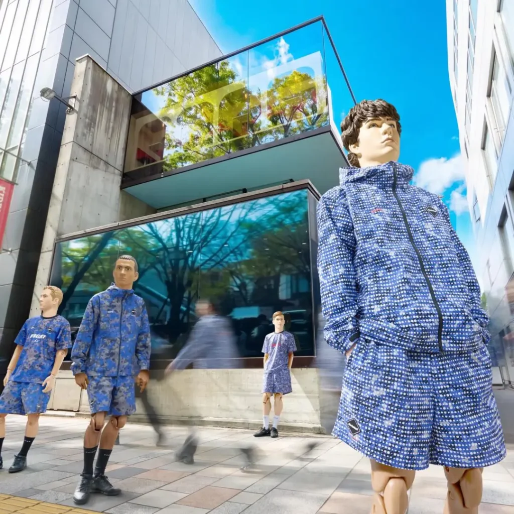 Futuristic streetwear presentation featuring mannequin figures dressed in coordinated blue patterned F.C. Real Bristol apparel outside a modern glass-and-concrete building under a bright sky. The collection includes lightweight jackets, shorts, and athletic sets covered in pixelated dot camouflage graphics, styled with black sneakers and sportswear silhouettes. Motion-blurred pedestrians move through the foreground, contrasting the static mannequins and emphasizing the campaign’s blend of football culture, urban architecture, and contemporary Japanese street fashion