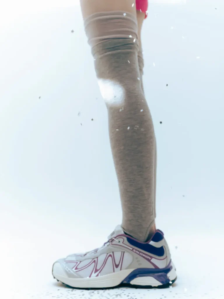 Side-profile editorial image of the Salomon x Ray BEAMS sneaker styled with sheer knee-high socks against a bright minimalist backdrop. The sneaker features silver mesh construction with flowing purple overlays, deep navy accents, and a sculpted trail-inspired sole, while scattered metallic star graphics enhance the campaign’s dreamy Y2K-meets-performance aesthetic