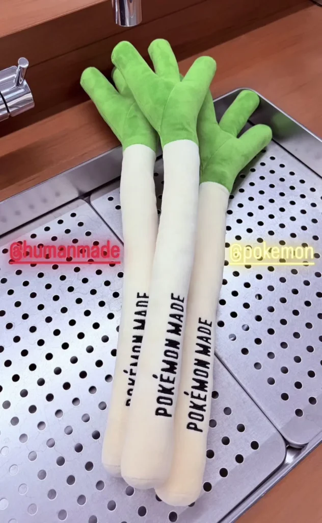 Three oversized plush green onion-inspired cushions from the Human Made x Pokémon collaboration lean across a perforated metallic surface, featuring soft lime-green tops, elongated cream stems, and bold “POKÉMON MADE” typography, staged against a warm wood and chrome interior with glowing @humanmade and @pokemon signage