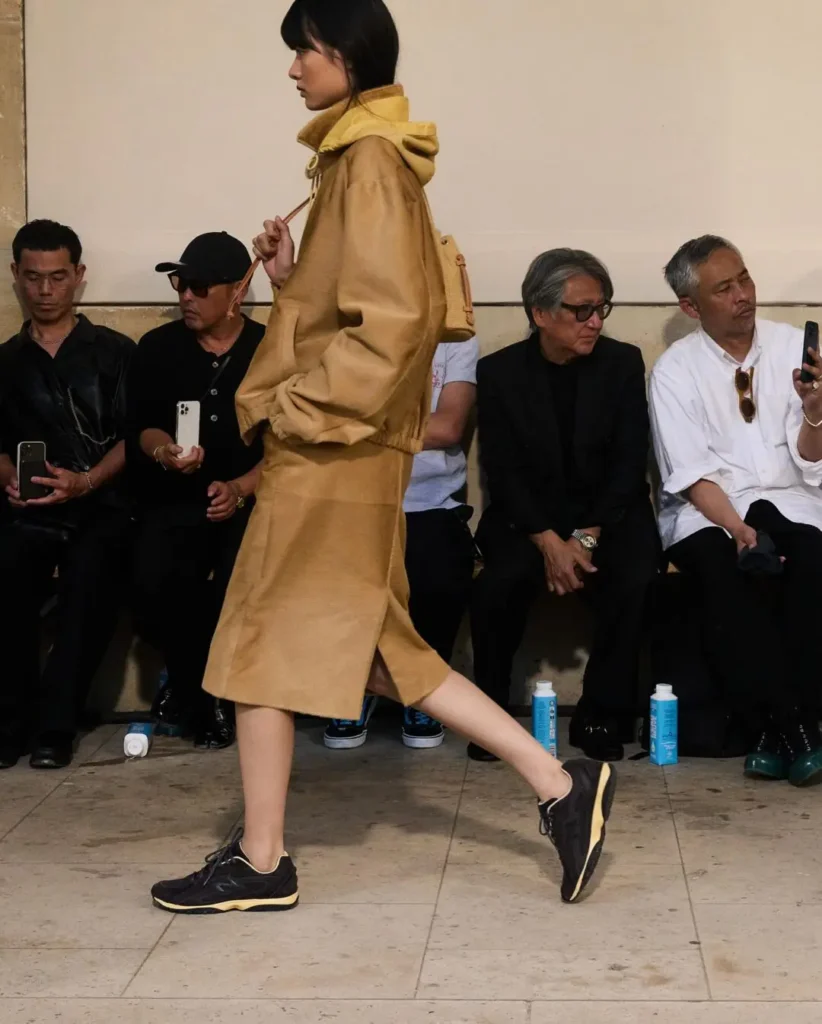A monochromatic black runner with sculpted tonal paneling and a softly yellowed midsole moves through the runway in understated fashion, pairing retro athletic influences with relaxed utilitarian tailoring. The streamlined silhouette reinforces the ongoing shift toward quieter, low-profile technical footwear within contemporary luxury styling