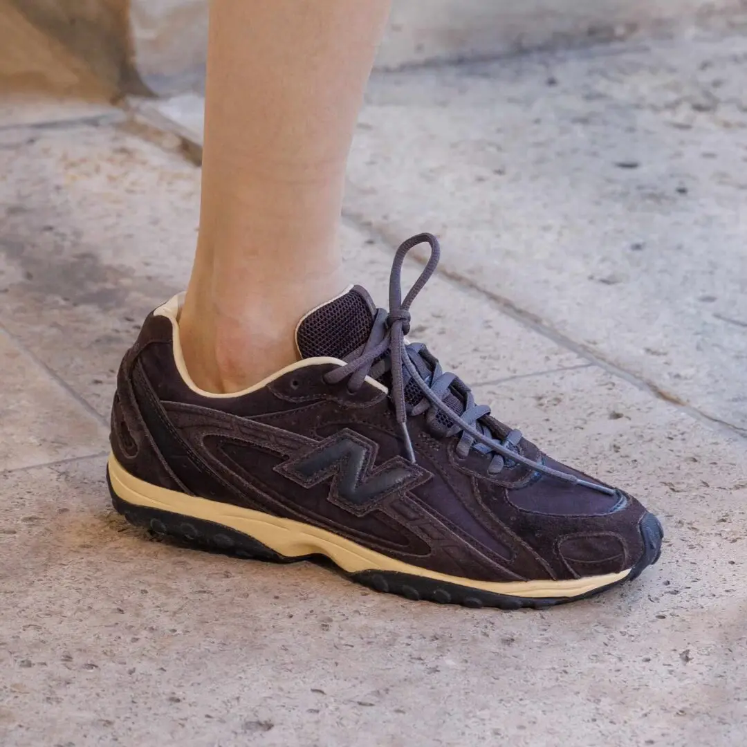 A dark tonal runner with layered suede and mesh construction pairs sculpted retro-running lines with a softly yellowed cream midsole, giving the silhouette an understated archival feel. The low-profile sneaker reflects the continued movement toward refined technical footwear rooted in early-2000s performance aesthetics