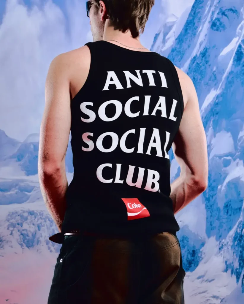 Campaign image showing the back of a fitted black Anti Social Social Club tank top featuring oversized stacked white branding and a small red Coca-Cola logo near the hem. Styled with dark denim and sunglasses against a snow-covered mountain backdrop, the look merges minimalist streetwear graphics with outdoor-inspired editorial imagery