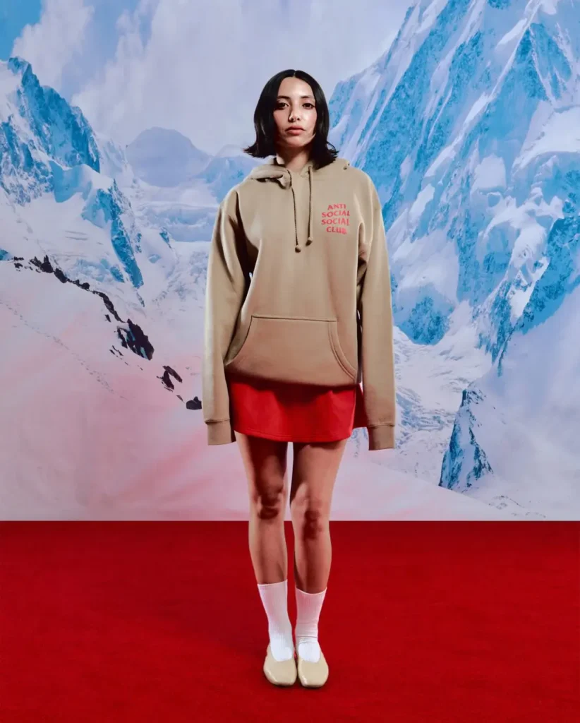 Editorial campaign portrait featuring a model wearing an oversized beige Anti Social Social Club hoodie styled over a vivid red mini skirt. Set against a dramatic snow-covered mountain backdrop and bold red studio floor, the minimal look combines muted outerwear tones with sharp color contrast, while the small chest logo adds subtle collaborative streetwear branding