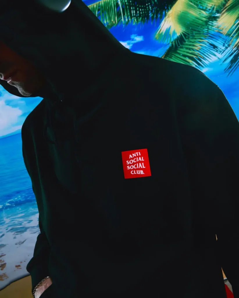 Moody campaign close-up featuring a model in a black Anti Social Social Club hoodie set against a saturated tropical beach backdrop with palm trees and ocean scenery. The minimalist garment is accented by a bold red square chest logo, while the shadowed hood styling and cinematic lighting create a darker, more atmospheric interpretation of the collaboration’s streetwear aesthetic