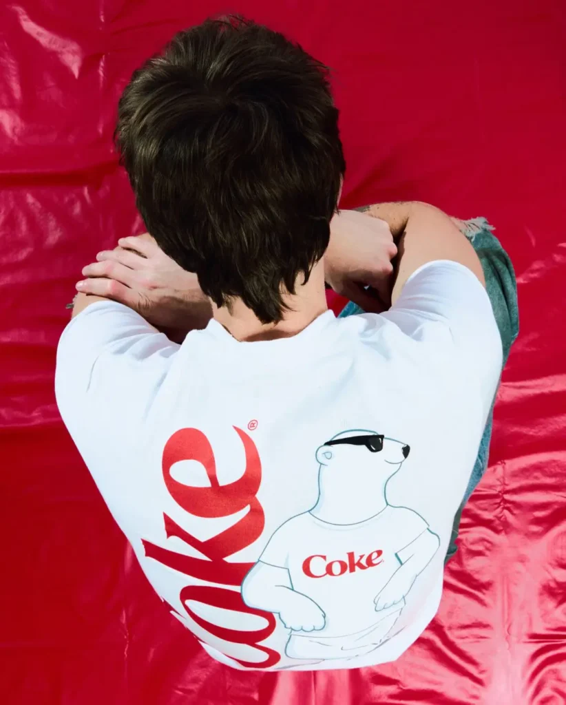 Overhead-style campaign image featuring the back graphic of a white Coca-Cola collaboration T-shirt against a vivid red backdrop. The design combines oversized red “Coke” lettering with a playful polar bear illustration wearing sunglasses and a Coca-Cola shirt, reinforcing the capsule’s nostalgic soda branding through a clean, graphic-heavy streetwear presentation