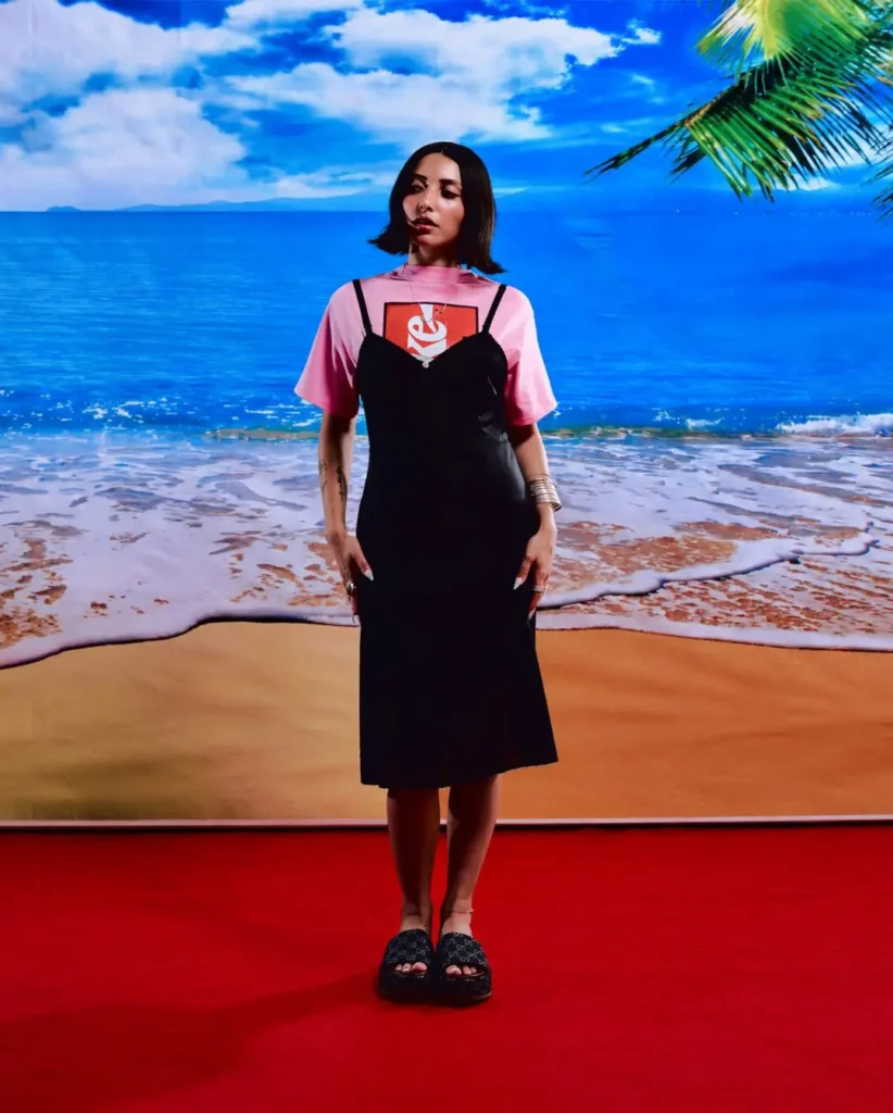 Full-body campaign portrait featuring a model styled in a black slip-style dress layered over a pastel pink Coca-Cola x Anti Social Social Club graphic T-shirt. Set against a vivid tropical beach backdrop with blue ocean water, palm trees, and a bold red studio floor, the look blends relaxed summer imagery with contemporary streetwear and minimalist fashion styling