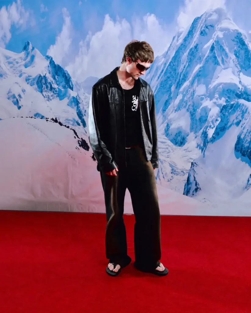 Full-body editorial campaign image featuring a model styled in a black leather jacket layered over a black Coca-Cola graphic top, paired with relaxed dark trousers, flip-flops, and oversized sunglasses. Positioned against a dramatic snow-covered mountain backdrop and vivid red floor, the look blends minimalist monochrome styling with subtle retro Coca-Cola branding and contemporary streetwear influences