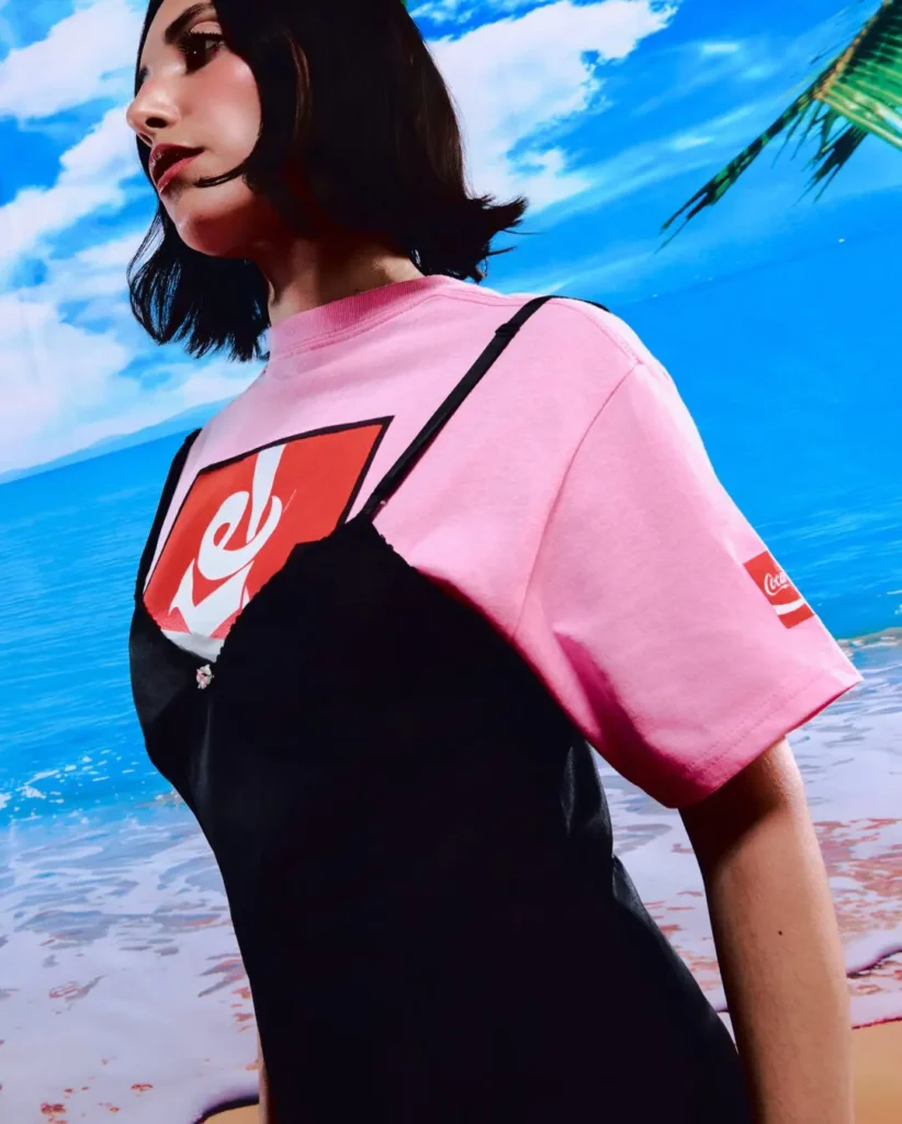 Fashion campaign image featuring a model wearing a pastel pink Coca-Cola x Anti Social Social Club graphic T-shirt layered beneath a black slip-style dress. The tropical backdrop of blue skies and ocean water amplifies the collection’s playful summer aesthetic, while Coca-Cola branding appears prominently across the chest graphic and sleeve patch