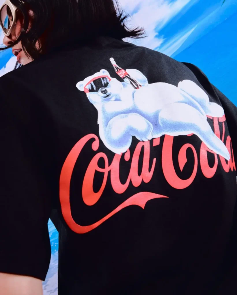 Close-up campaign image highlighting the back graphic of a black Coca-Cola collaboration T-shirt featuring the brand’s oversized red script logo beneath a lounging polar bear illustration wearing sunglasses and balancing a Coca-Cola bottle. Framed against a vivid tropical sky backdrop, the playful artwork channels retro soda advertising nostalgia through a contemporary streetwear presentation