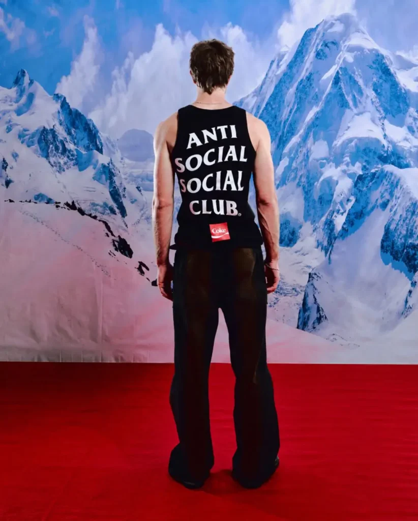 Full-body rear campaign portrait featuring a model wearing a fitted black Anti Social Social Club tank top with oversized stacked white branding and a small red Coca-Cola logo detail near the lower back. Styled with relaxed dark trousers against a dramatic snow-covered mountain backdrop and vivid red floor, the image blends minimalist streetwear graphics with stark alpine-inspired editorial scenery
