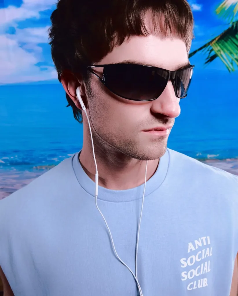 Close-up campaign portrait featuring a model in a pale blue sleeveless Anti Social Social Club top with stacked white chest branding, styled with oversized wraparound sunglasses and wired earphones. Set against a vivid tropical beach backdrop with blue ocean water and palm trees, the image channels an early-2000s-inspired resort aesthetic through minimalist streetwear styling