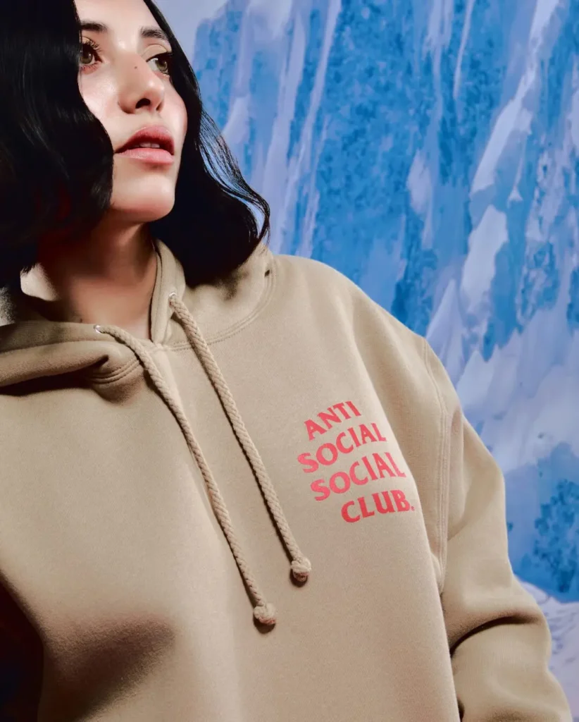 Close-up campaign portrait featuring a model wearing an oversized beige Anti Social Social Club hoodie with bold pink chest branding. Framed against an icy mountain backdrop, the neutral-toned garment contrasts with the vivid logo treatment, while the relaxed fit and soft color palette reinforce the collection’s understated streetwear aesthetic