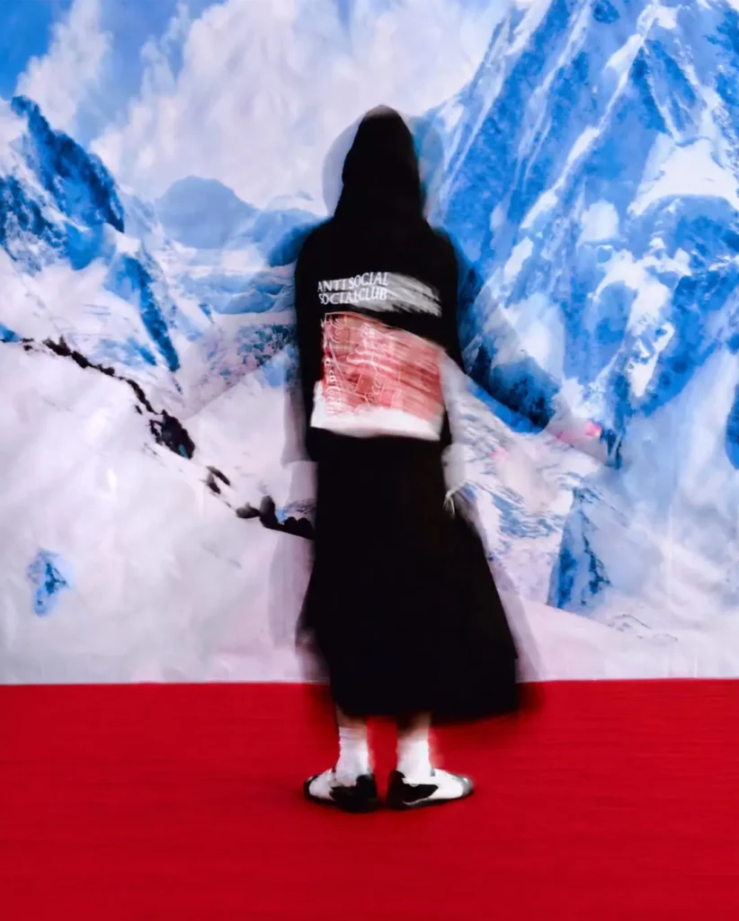 Blurred editorial campaign image featuring a model in a black Anti Social Social Club x Coca-Cola hoodie captured in motion against a dramatic icy mountain backdrop and vivid red floor. The distorted movement effect emphasizes the hoodie’s back graphic combining warped Anti Social Social Club branding with a red Coca-Cola can collage, creating a surreal, high-energy streetwear view