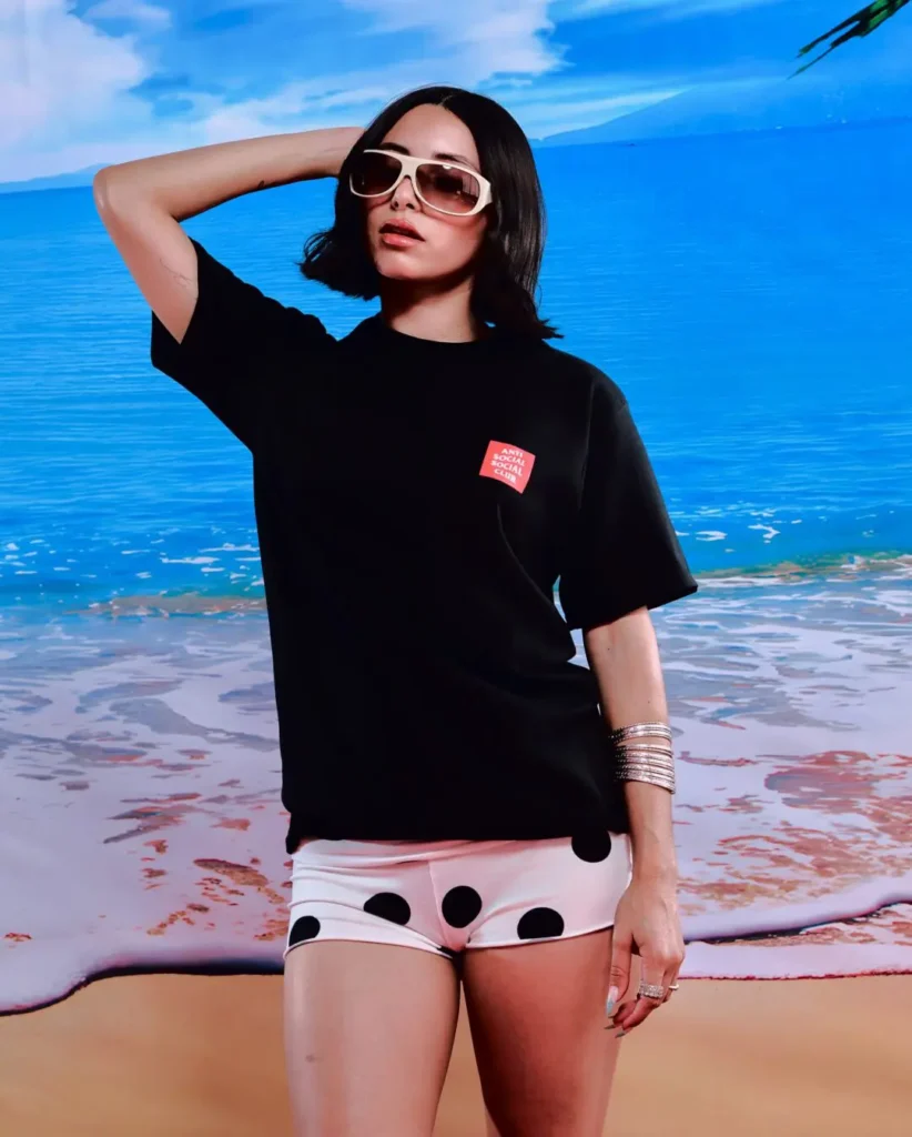 Campaign portrait featuring a model styled in an oversized black Anti Social Social Club T-shirt with a small red square chest logo, paired with playful black-and-white polka dot shorts. Oversized cream-framed sunglasses, stacked silver bangles, and a saturated tropical beach backdrop complete the look, blending retro vacation imagery with relaxed contemporary streetwear styling