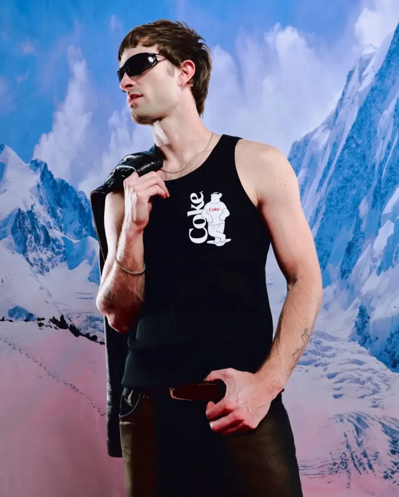 Editorial-style campaign portrait featuring a model in a fitted black tank top printed with Coca-Cola branding and the label’s signature polar bear mascot graphic across the chest. Styled with futuristic wraparound sunglasses and a leather jacket draped over the shoulder, the look contrasts sleek nightlife-inspired streetwear against a dramatic icy mountain backdrop, reinforcing the collection’s surreal blend of winter imagery and nostalgic soda iconography