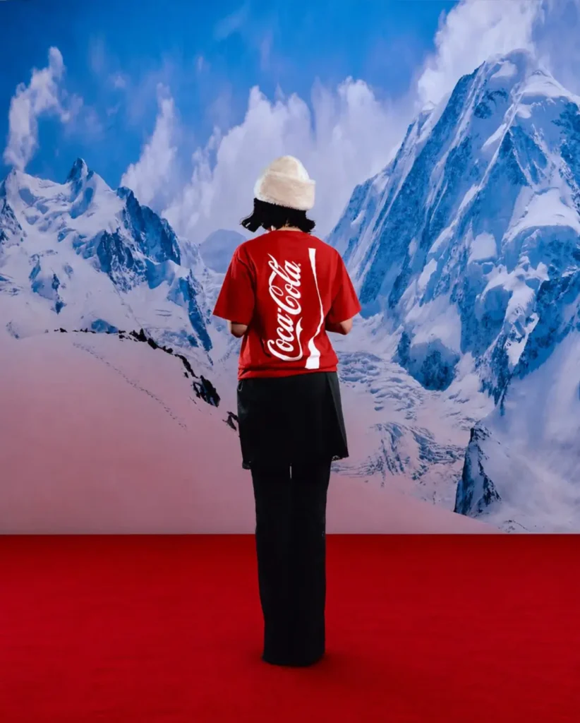 Rear-view editorial campaign image featuring a model in a vivid red Coca-Cola T-shirt with oversized vertical logo graphics and bottle-inspired white striping across the back. Styled with dark wide-leg trousers and a soft cream beanie against a towering snow-covered mountain backdrop, the composition blends nostalgic soda branding with cinematic winter-surrealist fashion imagery