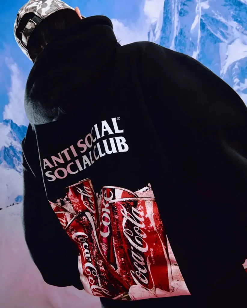 Detailed campaign close-up of a black hoodie featuring oversized Anti Social Social Club branding layered above a collage-style Coca-Cola can graphic bursting through ice textures. Shot from the back at an angled perspective, the image combines bold streetwear typography with classic soda iconography against a dramatic snow-covered mountain backdrop, emphasizing the collaboration’s cold-climate fantasy aesthetic