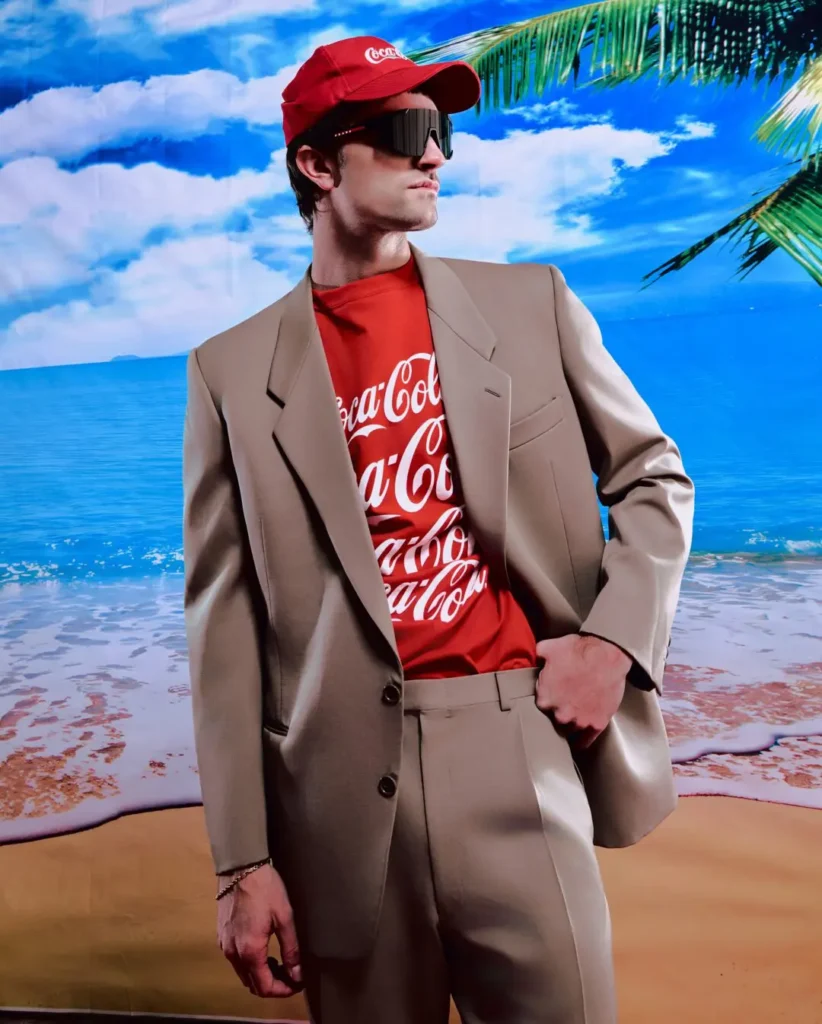 Editorial-style campaign portrait featuring a model in a relaxed beige tailored suit layered over a vivid red Coca-Cola x Anti Social Social Club graphic T-shirt. Styled with oversized futuristic sunglasses and a matching red Coca-Cola cap, the look contrasts formal tailoring with playful streetwear branding against a saturated tropical beach backdrop of palm trees, blue skies, and ocean water