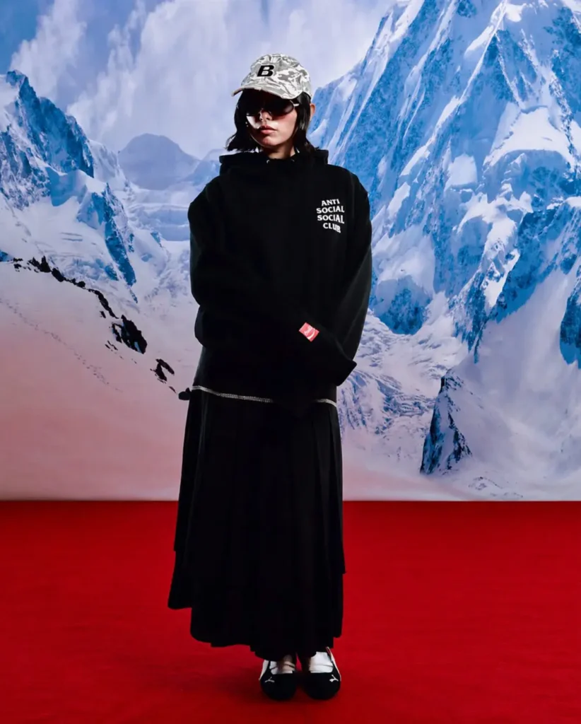 Studio-style campaign image featuring a model dressed in an oversized black Anti Social Social Club hoodie paired with a flowing black skirt, styled against a dramatic snow-covered mountain backdrop and vivid red floor. Accessories including a camouflage cap with a bold “B” logo and oversized dark sunglasses add an alpine-meets-streetwear atmosphere, while subtle Coca-Cola collaboration detailing appears on the sleeve tag