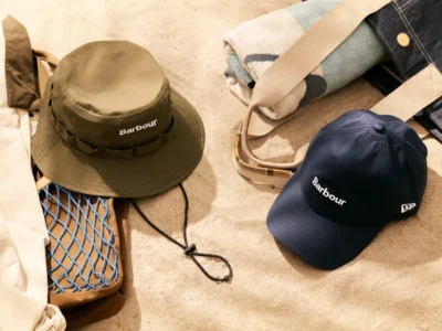 Flat-lay campaign image featuring the debut Barbour x New Era headwear capsule styled in an outdoor setting. An olive technical adventure hat with tonal webbing and a chin cord sits beside a navy performance cap featuring Barbour branding and subtle New Era embroidery. Surrounding accessories include a canvas tote, folded blanket, lightweight outerwear, and utility mesh detailing, reinforcing the collection’s heritage-meets-functional outdoor aesthetic