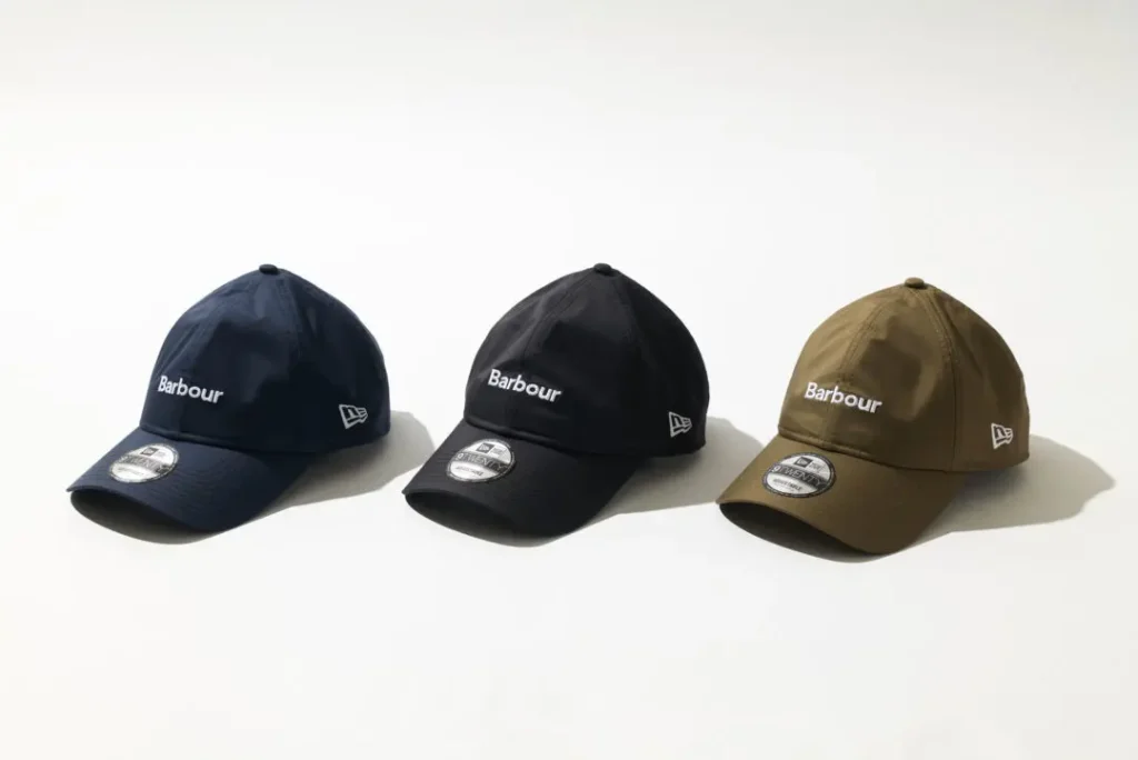 Studio product image of the Barbour x New Era 9TWENTY cap shown in three colorways—navy, black, and olive—arranged side by side against a clean white backdrop. Each cap features a smooth weather-resistant finish, curved brim, tonal panel construction, white Barbour wordmark at the front, and subtle New Era logo embroidery at the side, highlighting the collection’s understated utility-led approach