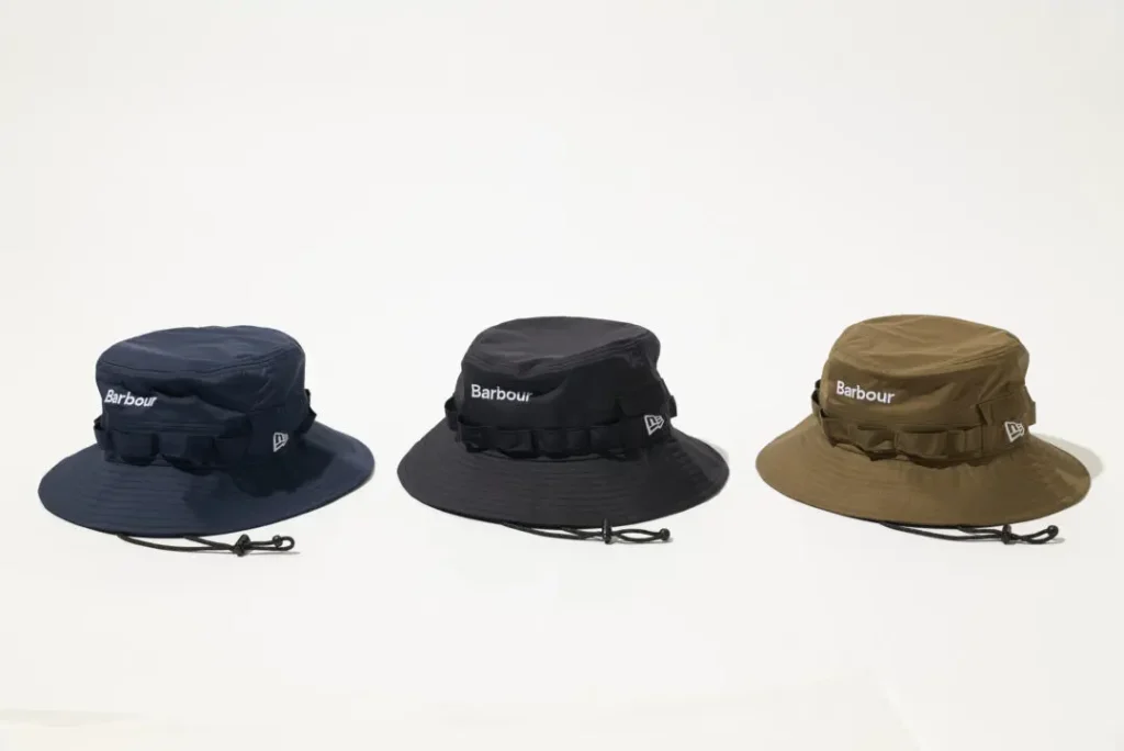 Studio product image showcasing the Barbour x New Era Adventure Hat in three colorways—navy, black, and olive—presented side by side against a clean white background. Each wide-brim silhouette features lightweight technical fabric, tonal utility webbing around the crown, an adjustable chin cord with toggle fastening, white Barbour branding at the front, and subtle New Era embroidery at the side, emphasizing the collection’s refined outdoor and weather-ready design language