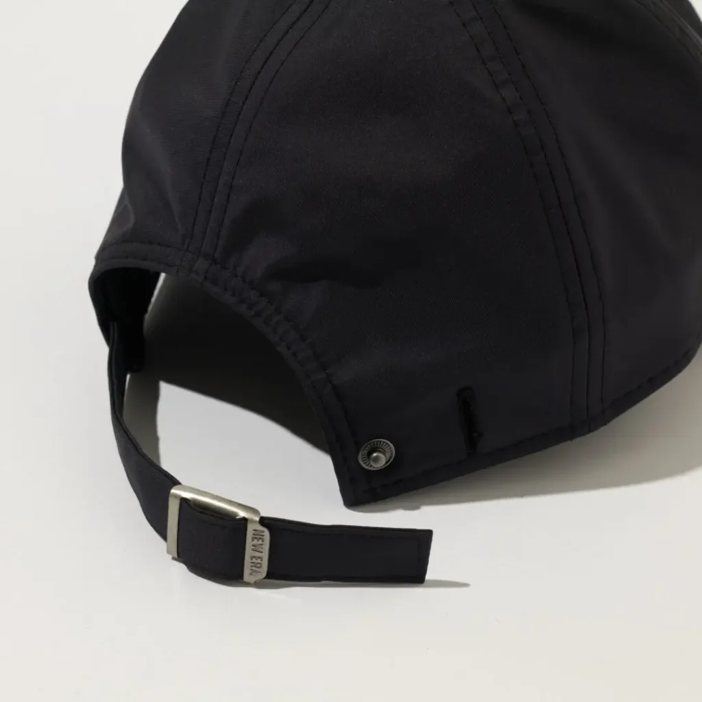 Close-up rear detail image of the Barbour x New Era 9TWENTY cap highlighting the adjustable back construction. The black technical fabric features a softly structured crown with tonal stitched panels, a curved rear opening, and a matching fabric strap secured by a brushed metal buckle engraved with New Era branding. Minimal hardware and clean finishing reinforce the collection’s refined outdoor utility and understated show aesthetic