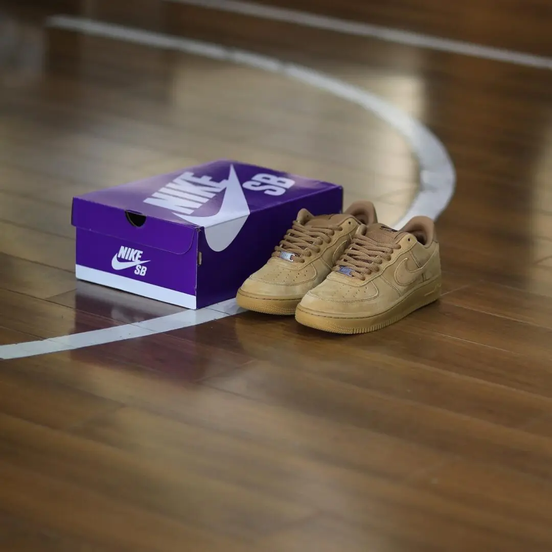 Pair of Nike SB Air Force 1 Low “Wheat” sneakers placed beside a purple Nike SB shoebox on a polished indoor basketball court, highlighting the premium suede construction and monochromatic gum-toned design