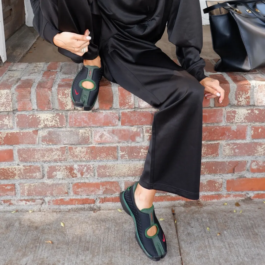 Lifestyle detail shot of a person seated on a brick ledge wearing dark green and black Nike Air Rift 2 sneakers with split-toe construction, open cutout detailing, and red Swoosh accents, styled with a relaxed black outfit and a structured black handbag in an outdoor setting