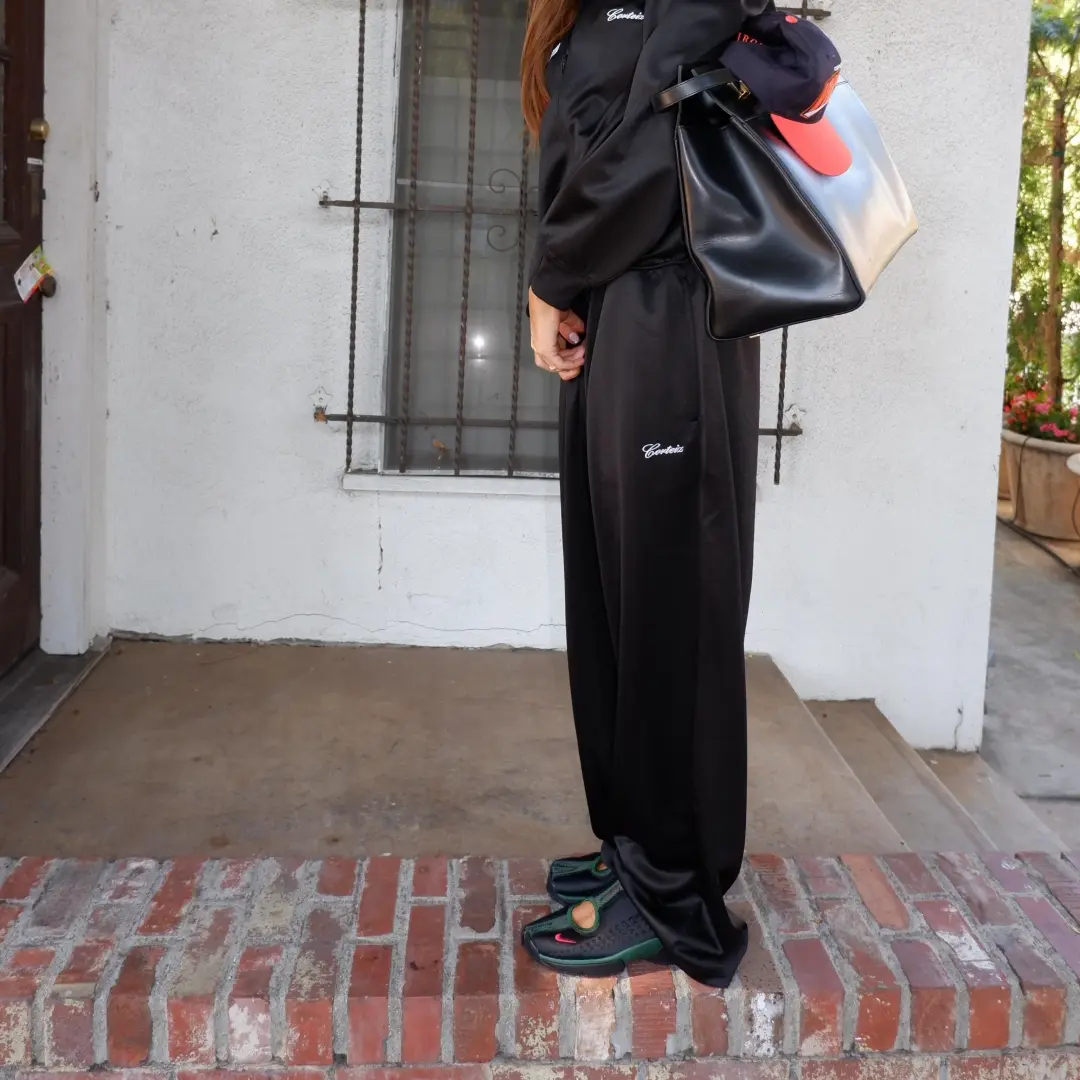 Full-length lifestyle image of a person standing on a brick step wearing dark green and black Nike Air Rift 2 sneakers with split-toe construction and red Swoosh accents, styled with oversized black track-inspired separates, a large structured black tote, and a layered casual accessory look against an outdoor residential backdrop