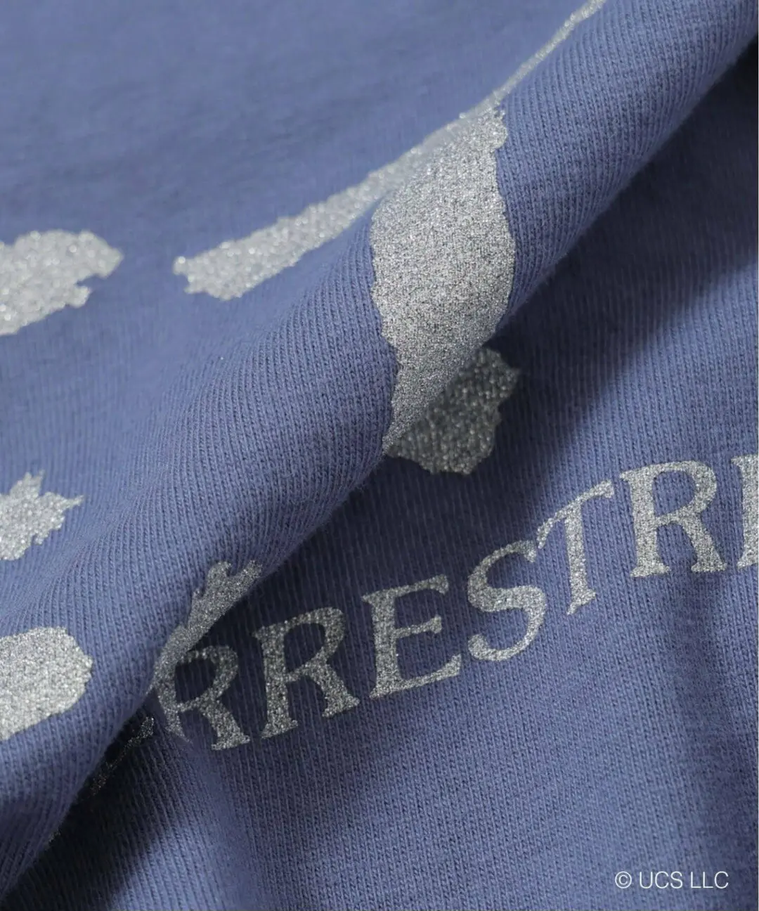 Close-up detail of the blue B.C STOCK × GOOD ROCK SPEED E.T. graphic T-shirt showing textured cotton jersey fabric, distressed silver-toned screen print, and vintage-inspired washed finish with partially visible “EXTRA-TERRESTRIAL” letter