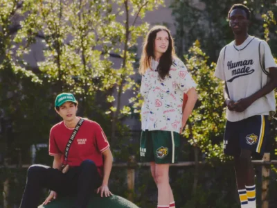 A sunlit outdoor campaign scene featuring three models styled in retro-inspired streetwear and varsity sportswear. One model wears a cream “Human Made” baseball jersey with matching athletic shorts and striped socks, while another pairs a white graphic short-sleeve shirt with green shorts featuring mascot-style patches. A third model sits casually in a red Human Made ringer tee, dark trousers, and a green branded cap with a crossbody strap. The setting’s warm natural lighting and tree-filled backdrop reinforce the collection’s relaxed vintage Americana and play collegiate aesthetic