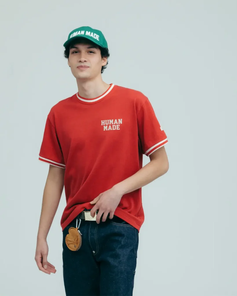 A minimalist studio campaign portrait featuring Human Made’s retro sportswear-inspired casual styling. The model wears a relaxed red ringer T-shirt with subtle Human Made chest branding and contrast striped trim, paired with dark raw denim and a bright green logo cap that adds a bold pop of color. A small baseball glove-inspired accessory clipped at the waist reinforces the collection’s nostalgic Americana and vintage athletic influences, while the clean neutral backdrop emphasizes the garment silhouettes and understated branding details