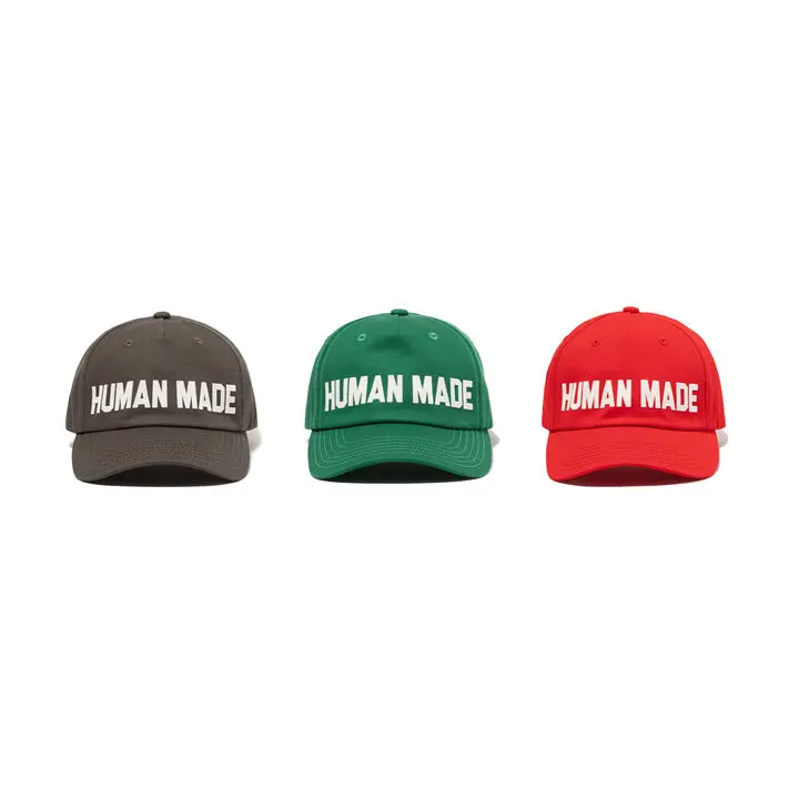 A clean product image displaying a trio of Human Made logo caps in charcoal grey, green, and bright red colorways against a neutral backdrop. Each structured baseball cap features bold embroidered Human Made lettering across the front panel, curved brims, and classic six-panel construction inspired by vintage athletic and workwear headwear. The minimalist presentation emphasizes the collection’s retro Americana influences, everyday utility, and signature balance between straightforward branding and timeless casual streetwear style