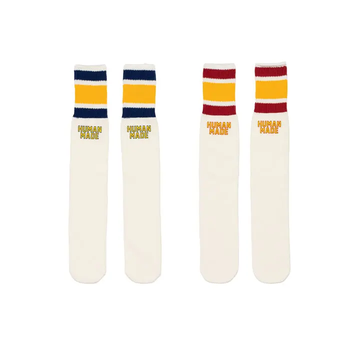 same A studio product image featuring two pairs of Human Made striped tube socks presented against a clean neutral backdrop. The retro athletic-inspired designs arrive in cream colorways accented with contrasting blue, yellow, and red varsity stripes near the cuff, alongside embroidered Human Made branding. Drawing heavily from vintage gym uniforms and classic Americana sportswear, the socks reinforce the collection’s nostalgic collegiate aesthetic while functioning as playful everyday styling accessories
