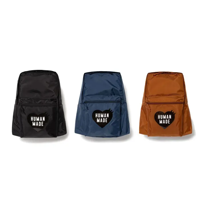 A product-focused studio image showcasing Human Made backpacks in black, navy, and brown colorways against a minimal neutral backdrop. Each lightweight utility-style backpack features a large front compartment and the brand’s signature heart logo patch prominently placed on the exterior pocket. Combining functional everyday design with nostalgic Americana-inspired branding, the accessories reflect Human Made’s balance of vintage sportswear influence, utilitarian practicality, and contemporary streetwear style