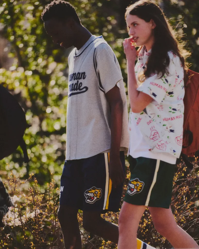 A candid outdoor campaign image showcasing Human Made’s blend of vintage athleticwear and playful graphic streetwear. One model wears a grey baseball-style jersey with script Human Made branding and navy athletic shorts featuring mascot-inspired patches and striped side detailing, while another pairs oversized green sports shorts with a white graphic button-up shirt covered in colorful doodle motifs and logos. Styled with backpacks and photographed in warm natural sunlight, the image captures the collection’s relaxed collegiate aesthetic and nostalgic Americana-inspired lifestyle direction