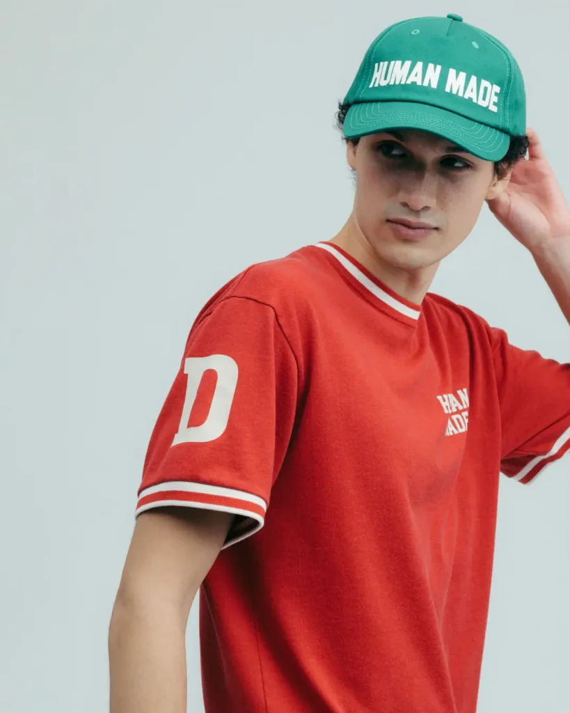 A clean studio-style campaign portrait featuring a model in Human Made’s retro athletic-inspired essentials. The look centers on a vivid red ringer T-shirt with contrast striped trim, varsity-style sleeve lettering, and subtle chest branding, paired with a bright green Human Made logo cap. Minimal styling and a neutral backdrop place emphasis on the garment’s vintage collegiate references, reinforcing the label’s signature blend of Americana sportswear nostalgia and contemporary streetwear simplicity