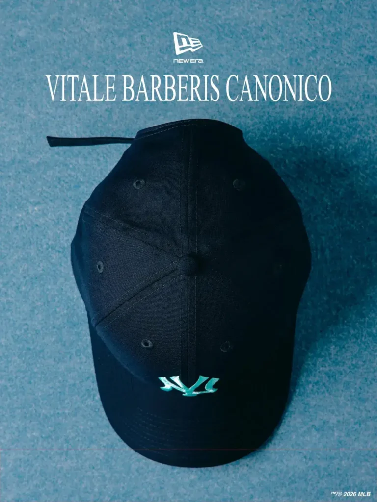 Top-down view of a black New Era 9FORTY cap rendered in premium Vitale Barberis Canonico wool with clean tonal paneling and a glowing Robin’s Egg Blue Yankees logo positioned near the visor edge. Set against a textured blue backdrop beneath bold collaborative branding, the image emphasizes understated tailoring influences blended with contemporary sportswear minimalism