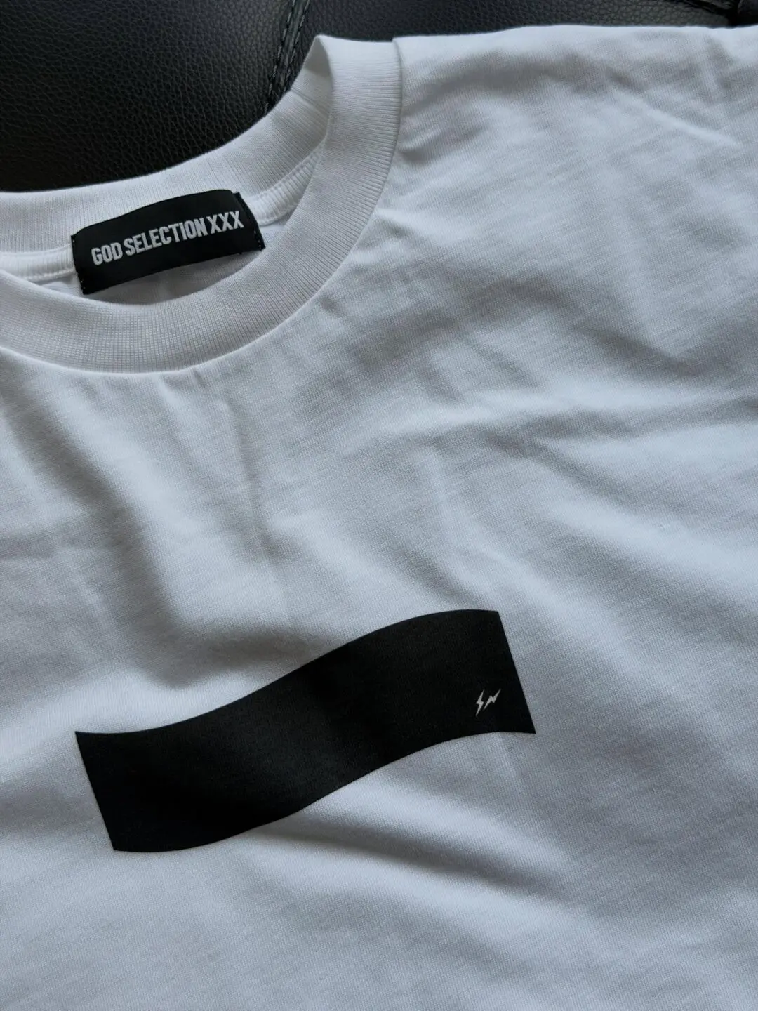 Close-up detail of a white crewneck T-shirt laid flat, featuring a minimalist black rectangular graphic across the chest with a small lightning-bolt style emblem near the edge. The neckline includes a black woven label reading “GOD SELECTION XXX,” emphasizing the brand’s clean monochrome aesthetic and premium streetwear presentation