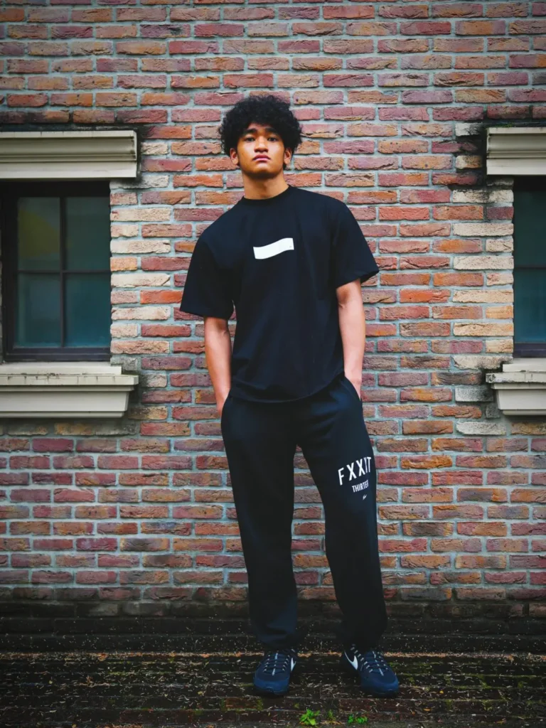 Full-body streetwear editorial portrait showing a person standing confidently against a textured red brick wall framed by two windows. The look features a monochrome black outfit consisting of an oversized graphic T-shirt with a minimalist white chest motif and matching relaxed sweatpants with bold white typography near the thigh. Hands-in-pocket styling, dark sneakers, and moody lighting create a clean, understated campaign aesthetic with a contemporary urban feel
