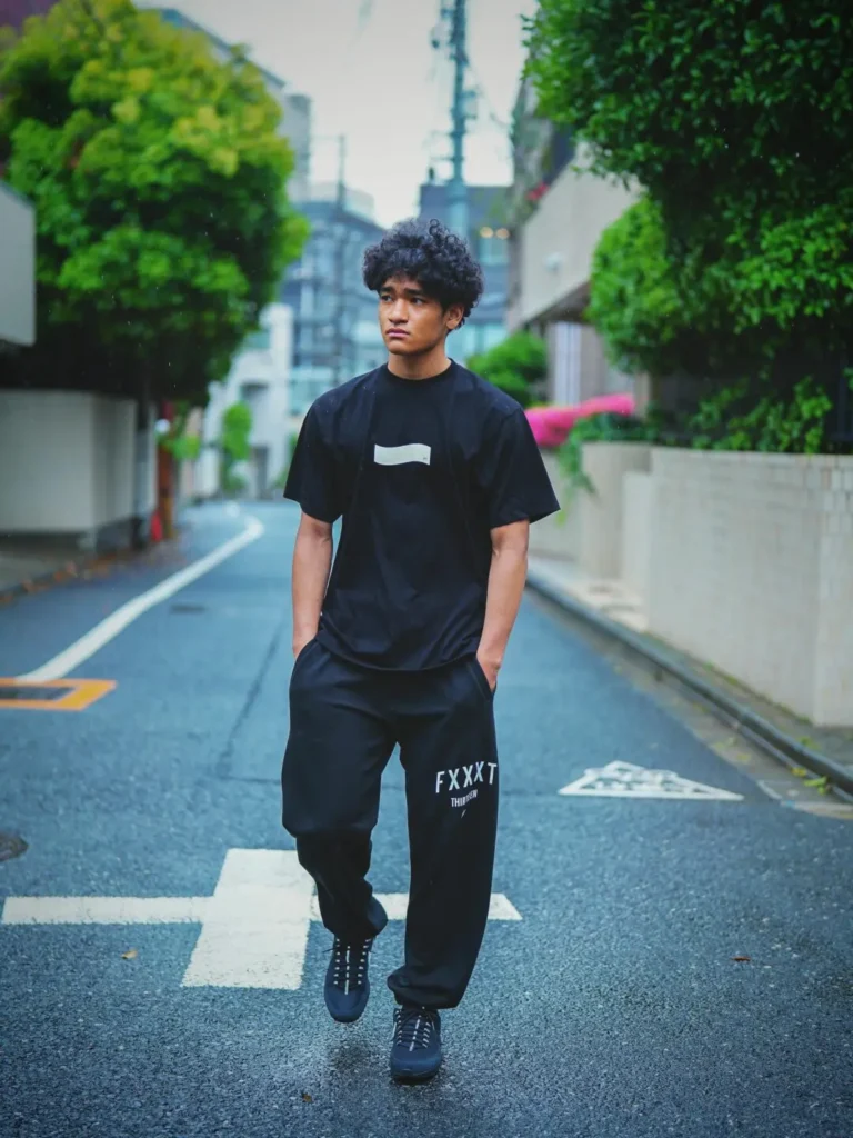 Streetwear editorial image showing a person walking through a quiet, rain-darkened city street while wearing a monochrome black set consisting of an oversized graphic T-shirt with a minimalist white chest motif and relaxed sweatpants featuring bold white typography near the thigh. With hands in pockets and a thoughtful side glance, the subject is framed by lush greenery and softly blurred urban architecture, creating a cinematic, understated campaign atmosphere