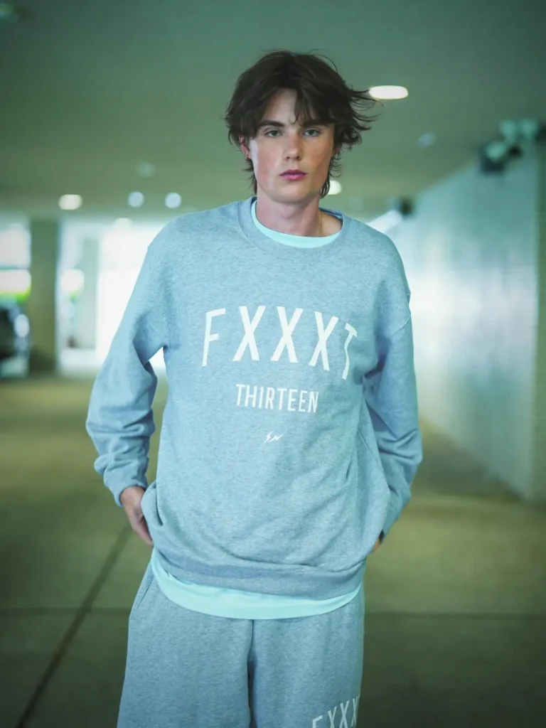 Streetwear editorial portrait showing a person standing in a softly lit indoor passage or parking space wearing a coordinated heather grey sweatshirt and sweatpants set. The oversized crewneck features bold white typography across the chest with smaller text and a subtle lightning-bolt style emblem beneath. With hands tucked casually into pockets and muted cinematic lighting, the image creates a relaxed, contemporary campaign aesthetic with an elevated athleisure feel