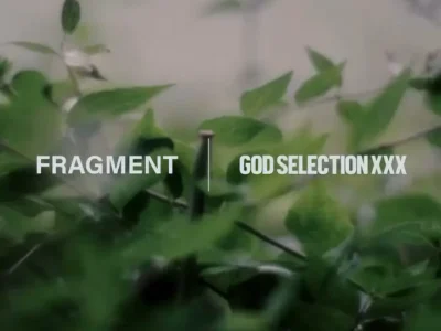 Soft-focus campaign visual featuring blurred green foliage in the foreground and a muted natural backdrop. Centered across the image are the wordmarks “FRAGMENT” and “GOD SELECTION XXX,” separated by a thin vertical line, creating a minimalist collaborative identity with a calm, atmospheric, and understated streetwear aesthetic