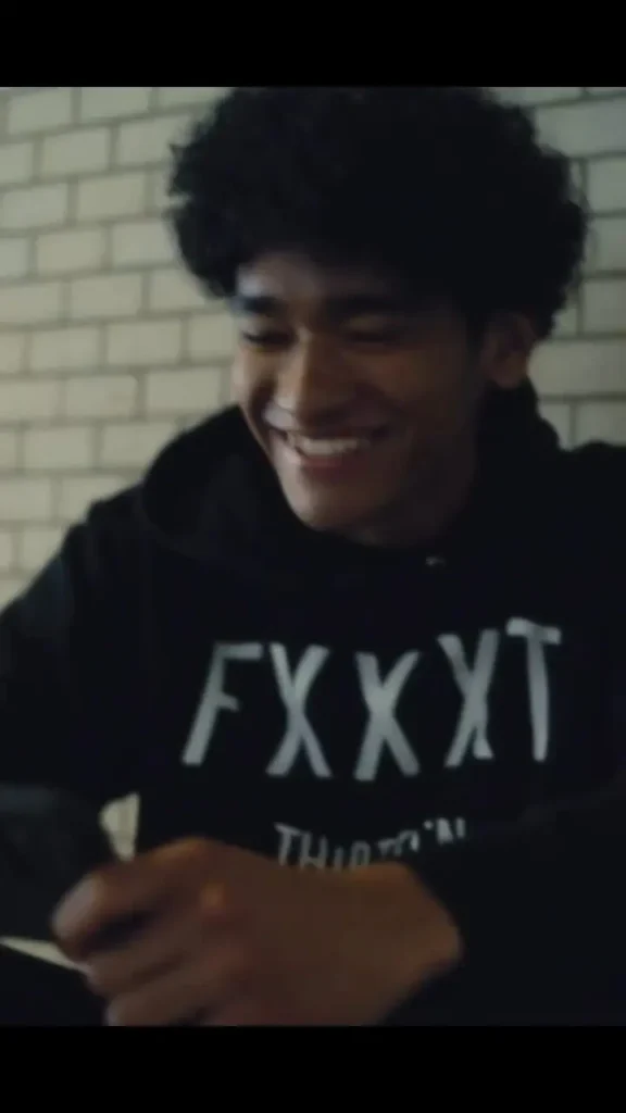 Soft-focus lifestyle campaign image showing a smiling person seated indoors against a light brick wall, wearing a black hoodie with bold white typography across the chest. The candid moment and muted lighting create an intimate, relaxed atmosphere that aligns with contemporary streetwear editorial aesthetics