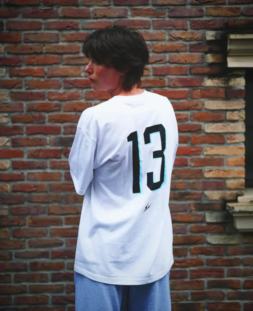 Streetwear editorial portrait showing a person standing with their back turned against a textured red brick wall while glancing over their shoulder. They wear an oversized white T-shirt featuring a large black “13” graphic across the back with a small lightning-bolt style emblem below, paired with relaxed grey sweatpants. The muted color palette and soft-focus treatment create a contemporary, understated campaign aesthetic with a casual urban mood